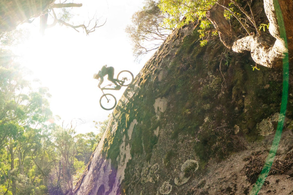 Biker riding a mountain bike through a rocky forest landscape with sunlight filtering through.